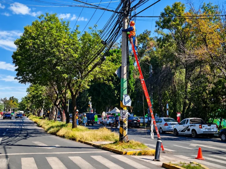 Trabajadores realizan labores sobre un poste con cableado aéreo en una vialidad urbana, como ejemplo de infraestructura de telecomunicaciones.